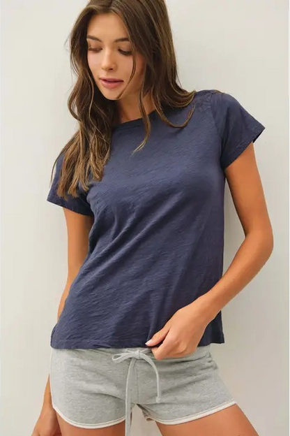 A woman wearing a short-sleeve crewneck t-shirt in a heathered navy blue color, paired with light gray drawstring shorts, standing against a plain light gray wall.