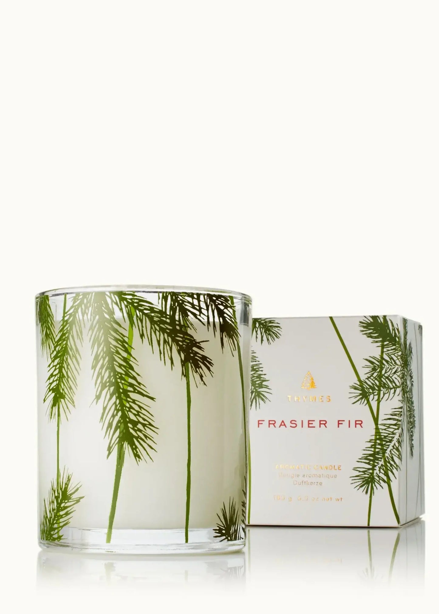 A glass jar candle with green fraser fir tree illustrations on a white wax base, shown next to its white packaging box featuring the same green pine design and gold text.
