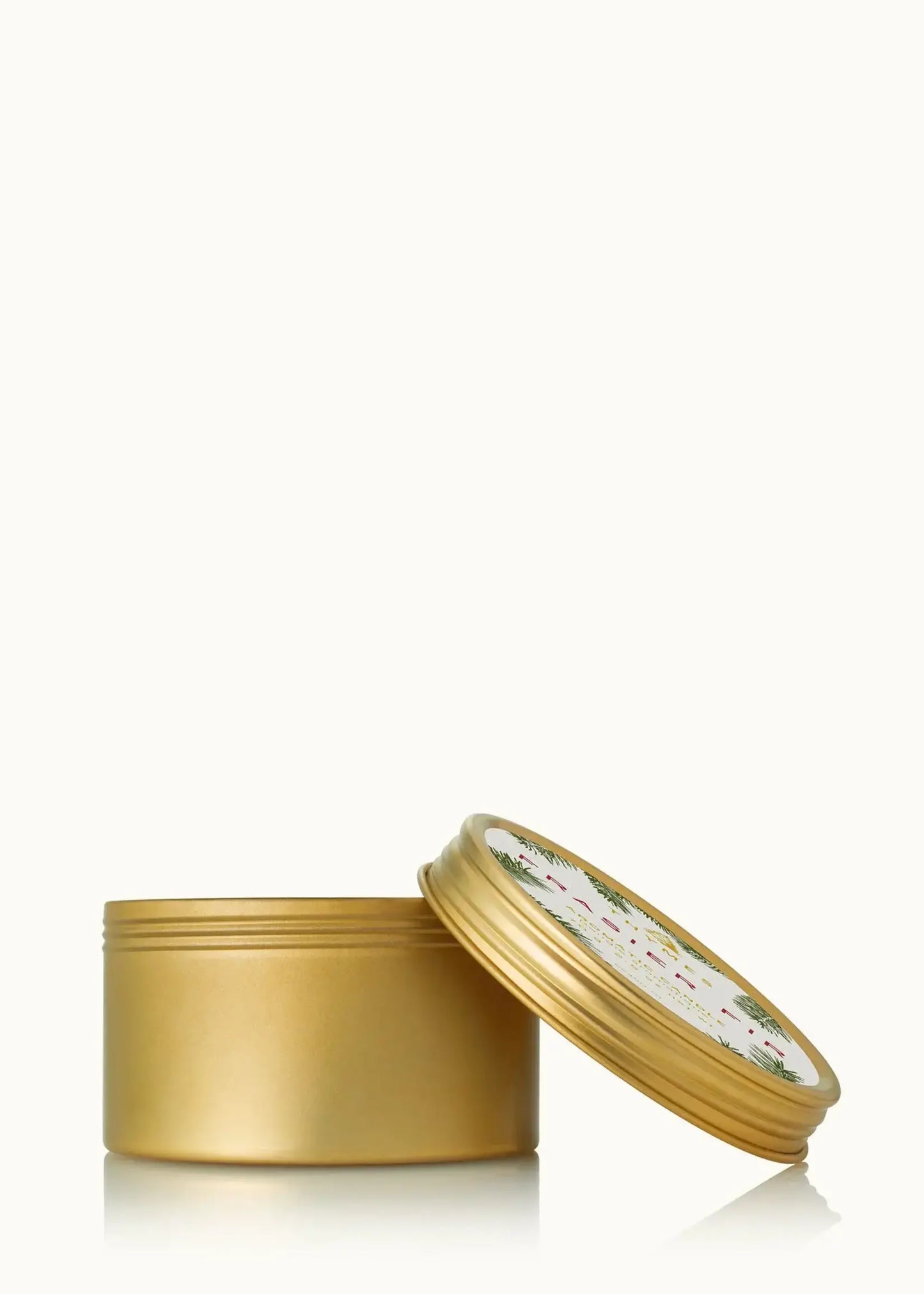 A gold metal tin container with its lid propped open, showing a white patterned label with green and red text, set against a plain white background.