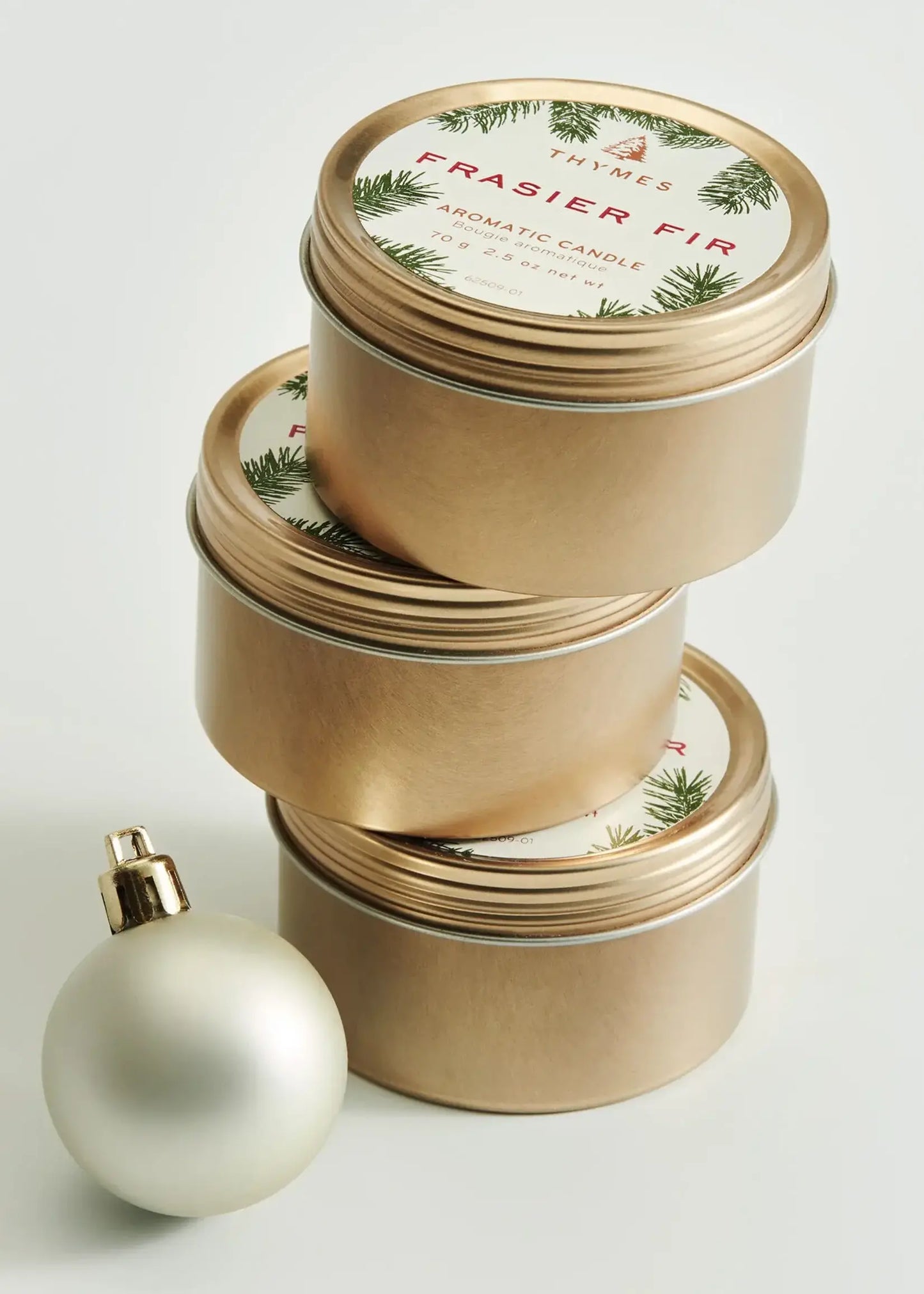 Three stacked round gold metal candle tins with white holiday labels reading 'Frasier Fir' and pine green illustrations, alongside a white spherical ornament on a clean white background.