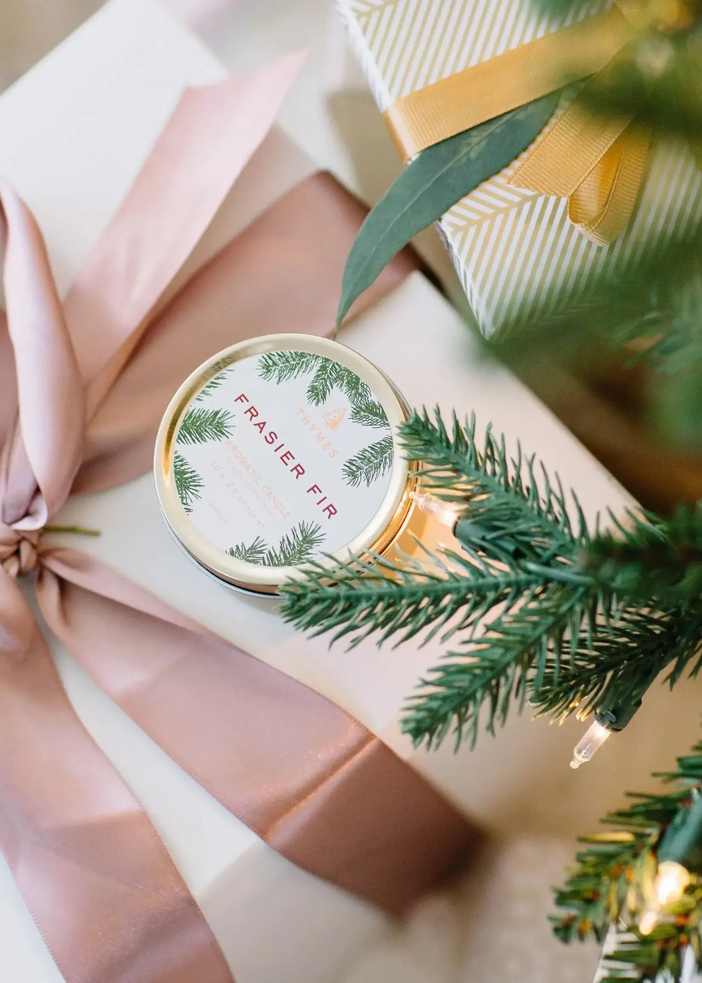 A round tin candle labeled 'FRAASIER PIR' with green pine illustrations on the lid, styled on a gift box with a blush pink ribbon, green pine branches, and a striped gift box.