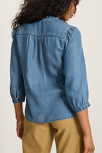 A woman shown from the back wearing a light blue denim button-up shirt with gathered puff sleeves and tan pants, photographed against a plain light beige studio background.