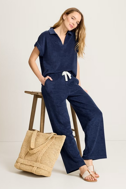 Woman in navy outfit sitting on a stool with a woven bag beside her on a light background