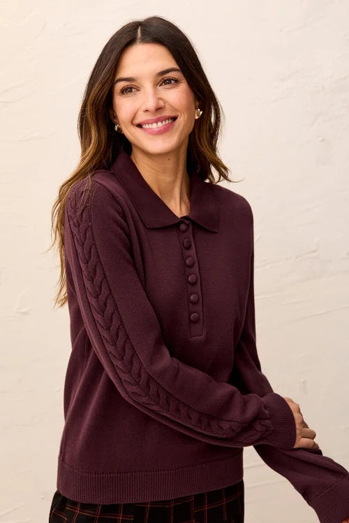 A woman wearing a dark plum purple knit polo sweater with cable and button details on the sleeves, standing against a light textured wall.