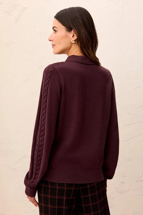 A woman shown from the back wearing a dark plum purple knit sweater with textured cable detailing on the sleeves, paired with dark plaid pants, against a textured off-white wall.
