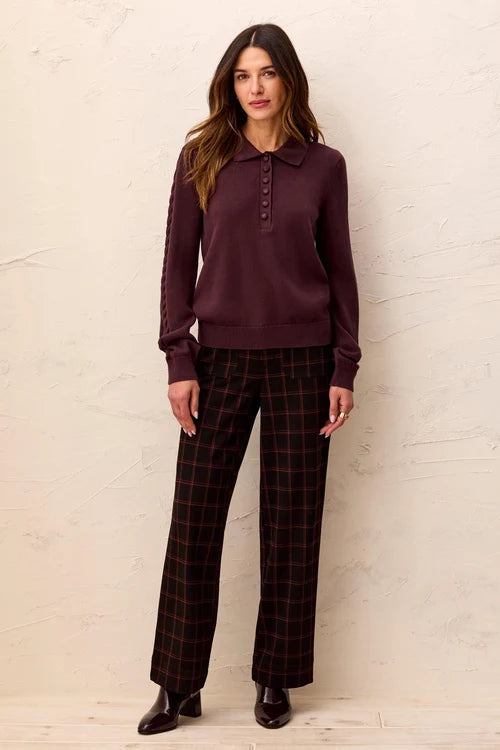 A woman modeling a dark plum long-sleeve polo knit top paired with dark red and black plaid trousers, standing against a textured off-white wall.