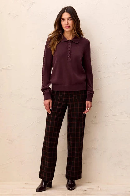 A woman modeling a dark plum long-sleeve polo knit top paired with dark red and black plaid trousers, standing against a textured off-white wall.
