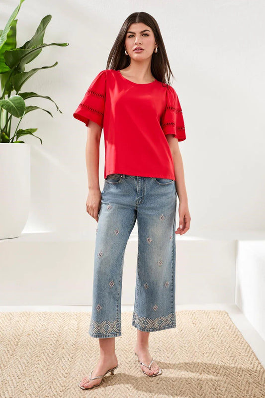 Woman wearing a red top and blue jeans standing indoors with a plant in the background
