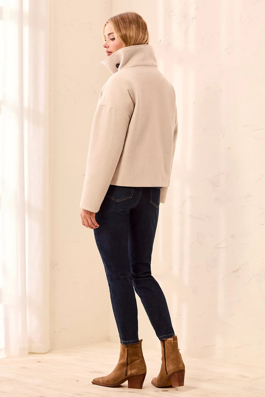 A woman wearing a beige cropped turtleneck jacket, dark blue skinny jeans, and brown ankle boots, standing in profile against a bright, softly lit cream-colored wall.