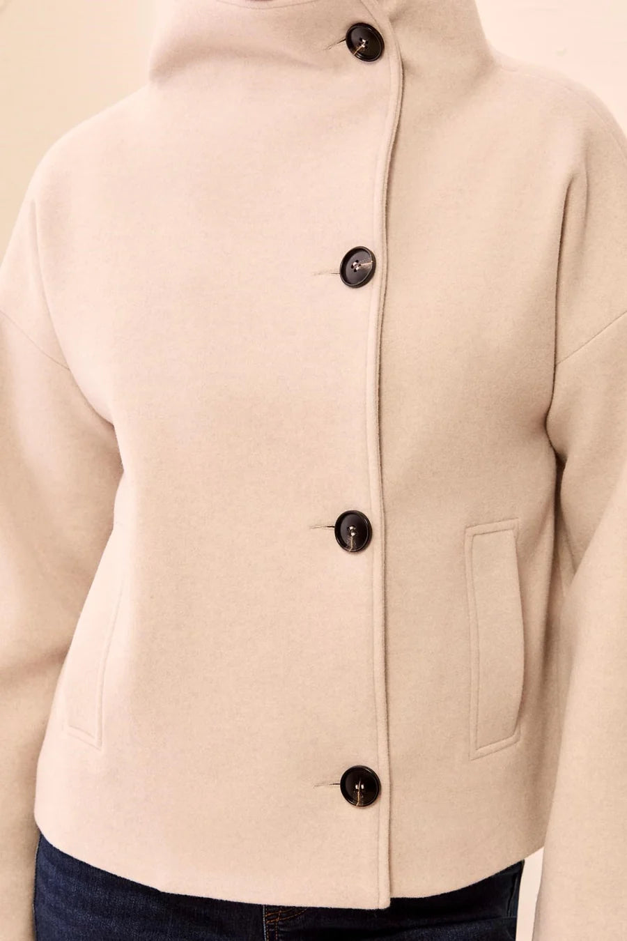 A close-up of a cream-colored cropped jacket with a high stand collar and four dark buttons, worn over dark blue jeans.