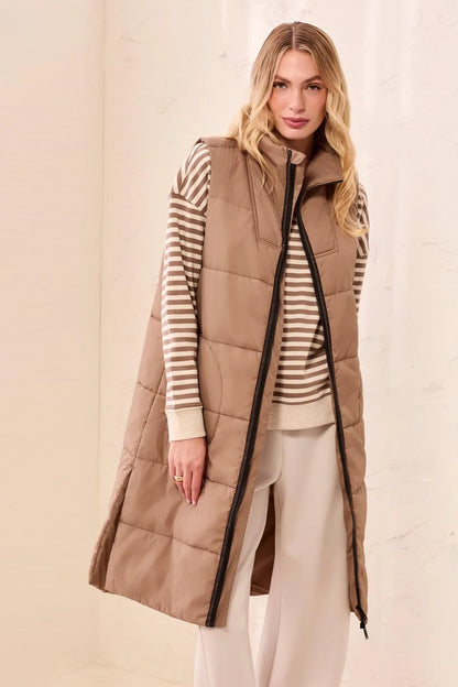 A woman modeling a long taupe-brown puffer vest over a beige and white striped sweater and cream pants, standing against a light beige textured wall.
