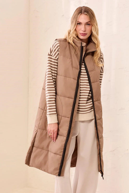 A woman modeling a long taupe-brown puffer vest over a beige and white striped sweater and cream pants, standing against a light beige textured wall.