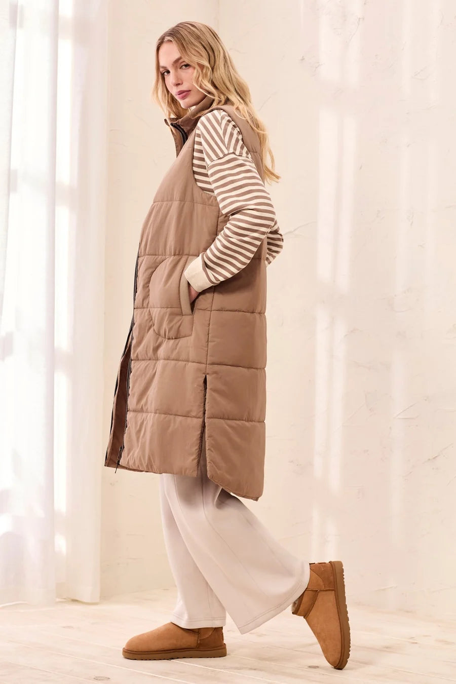 A woman wearing a long taupe-brown puffer vest over a beige and white striped sweater, white wide-leg pants, and tan slip-on boots, standing in a bright indoor setting with a textured cream wall.