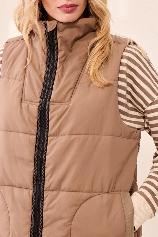 A close-up of a woman wearing a tan quilted puffer vest over a beige and brown striped long-sleeve shirt. The vest features a high collar and a black front zipper.