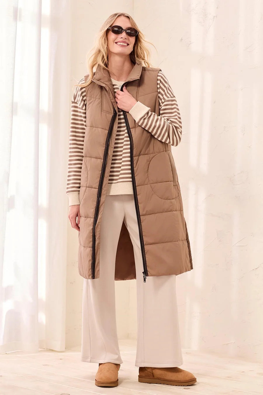 A woman modeling a long tan quilted puffer vest over a beige and white striped long-sleeve shirt, cream wide-leg pants, and tan slippers in a bright indoor setting with soft window light.