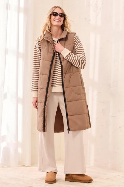A woman modeling a long tan quilted puffer vest over a beige and white striped long-sleeve shirt, cream wide-leg pants, and tan slippers in a bright indoor setting with soft window light.