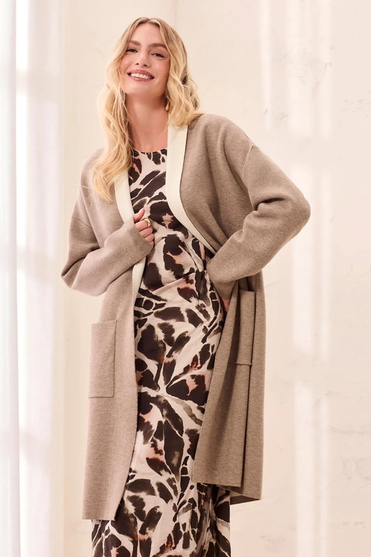 A woman wearing a long taupe knit cardigan over a brown and beige animal print dress, standing in a brightly lit indoor setting with a soft white background.