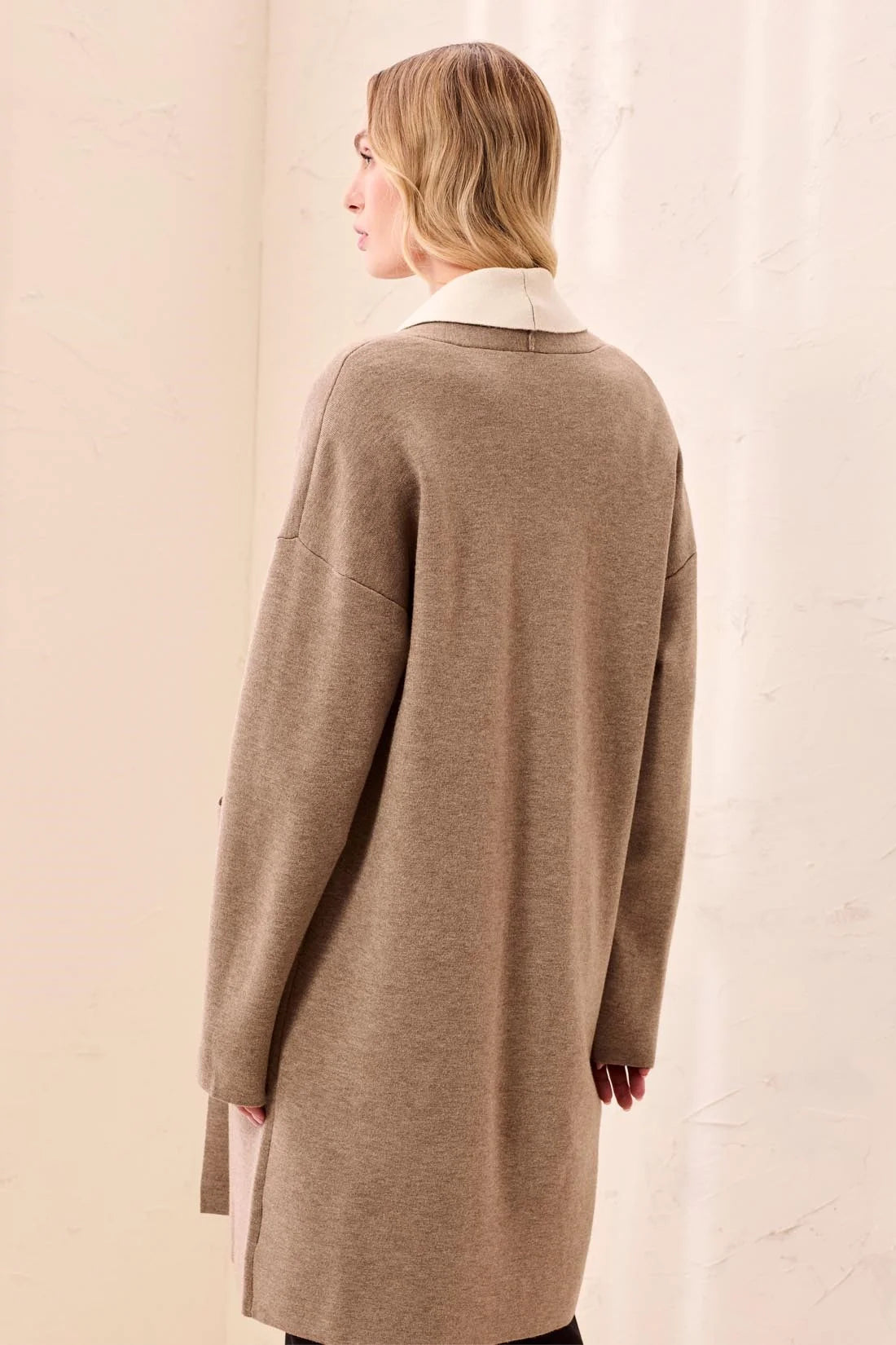 A woman shown from the back wearing a long, oversized taupe knit cardigan with a contrasting cream collar, set against a soft beige background.