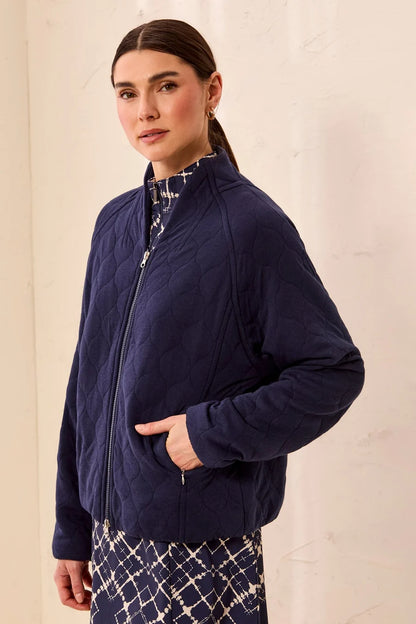 A woman wearing a navy blue quilted zip-up jacket over a patterned top and matching patterned skirt, standing against a light beige textured wall.