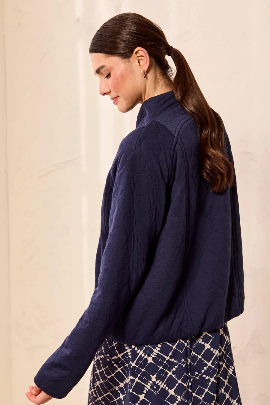 A woman wearing a dark navy blue textured long-sleeve mock-neck top and a patterned skirt, shown from a side-back angle against a light beige wall.