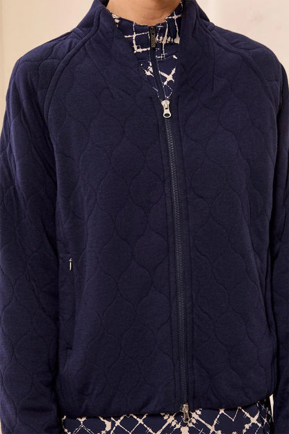 A close-up of a person wearing a navy quilted zip-up jacket over a patterned top. The jacket has a diamond-quilted texture, a full front zipper, and a stand collar.