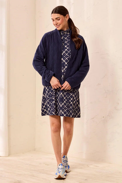 A woman modeling a navy blue zip-up fleece jacket over a navy and white patterned dress, standing in a minimalist cream-colored room with light wood floors.