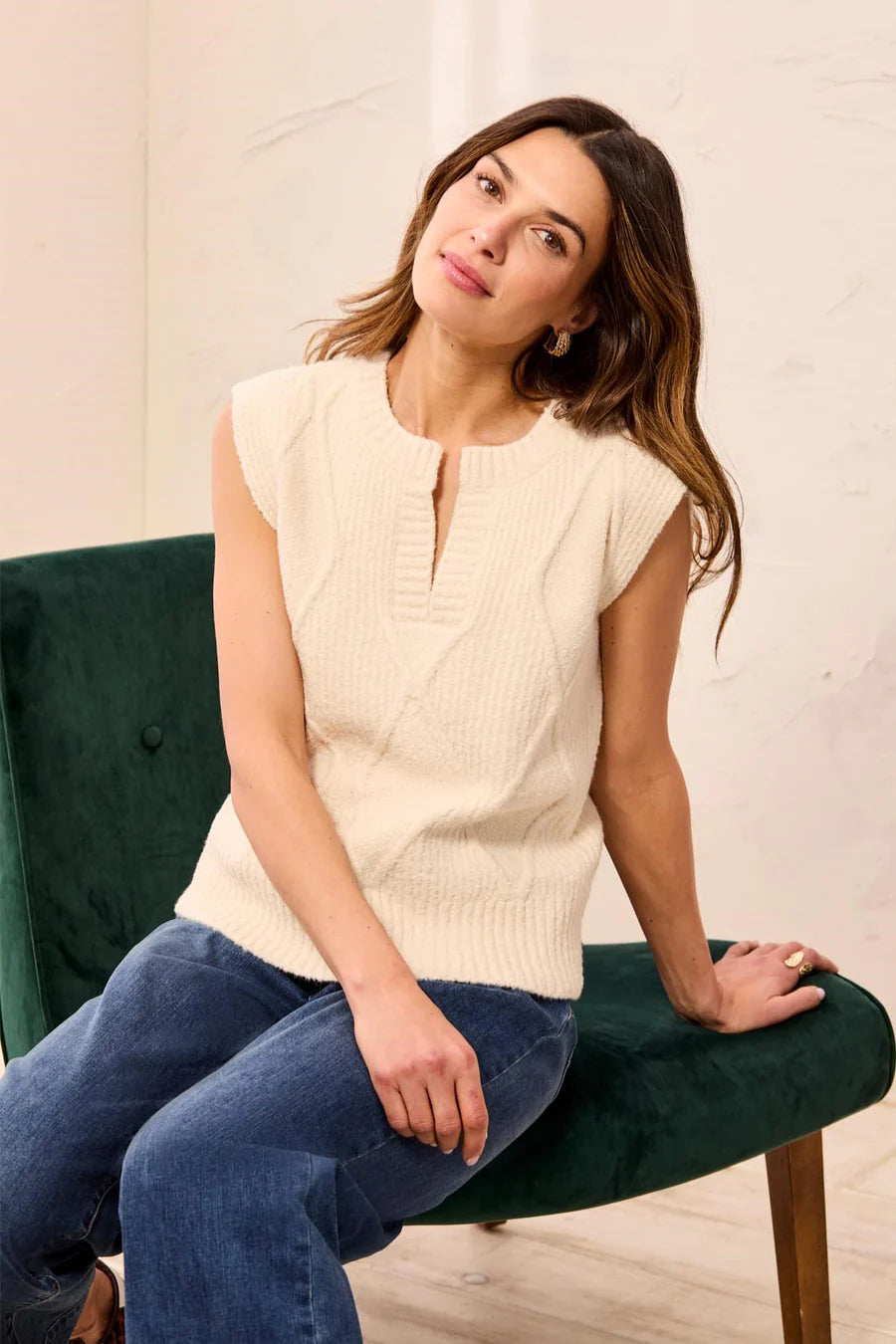 A woman wearing a cream ribbed knit sleeveless top and blue jeans sits on a dark green velvet armchair in a bright indoor setting.
