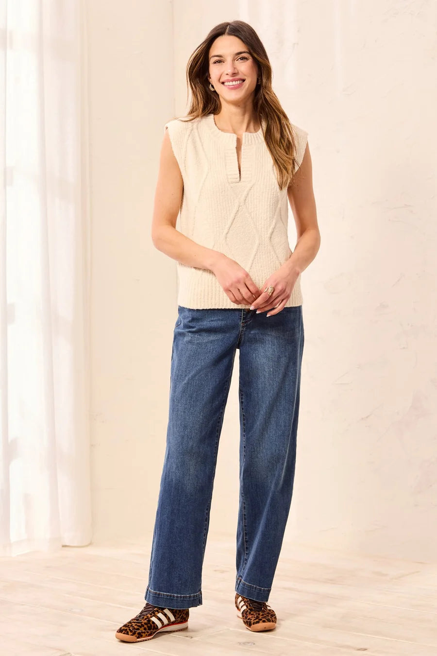 A woman modeling a cream sleeveless knit top with a split neckline and diamond cable-knit pattern, paired with blue jeans and leopard-print flat shoes, standing in a bright indoor setting.