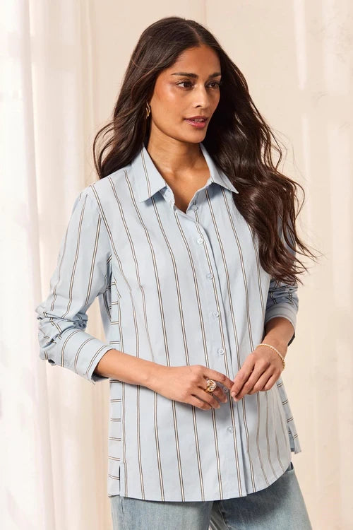 A woman wearing a light blue button-up shirt with thin vertical brown stripes, paired with light-wash jeans, standing against a soft beige curtain backdrop.