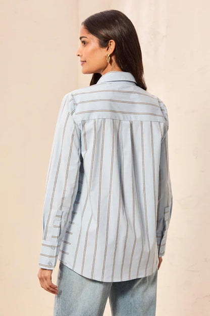 A woman shown from the back wearing a light blue striped button-up shirt and light-wash jeans, standing against a plain beige wall.
