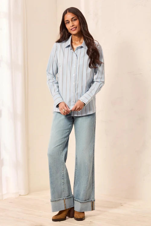 A woman modeling a light blue and white vertically striped button-down shirt paired with wide-leg light-wash jeans and brown ankle boots, standing in a softly lit indoor setting with a cream wall and white curtain.