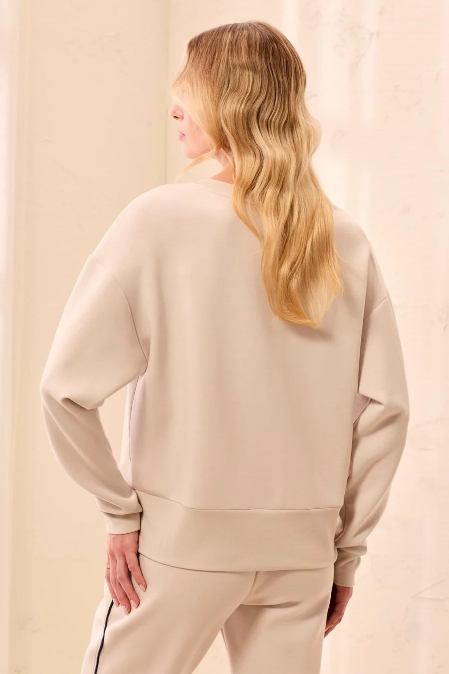A woman with long blonde hair is shown from behind wearing a light beige crewneck sweatshirt and matching sweatpants against a soft beige background.