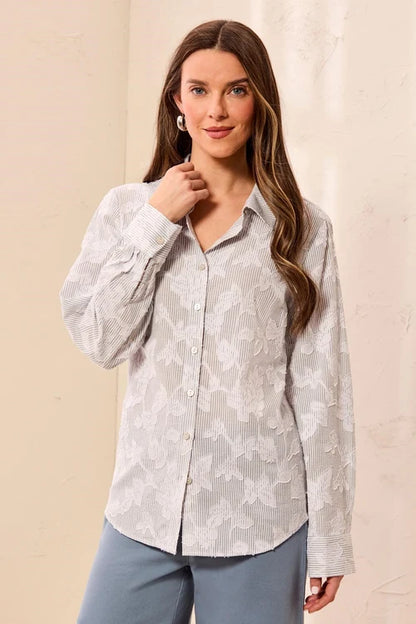 A woman modeling a light gray and white vertically striped button-down shirt with a subtle tonal floral jacquard pattern, paired with slate blue pants against a neutral beige background.