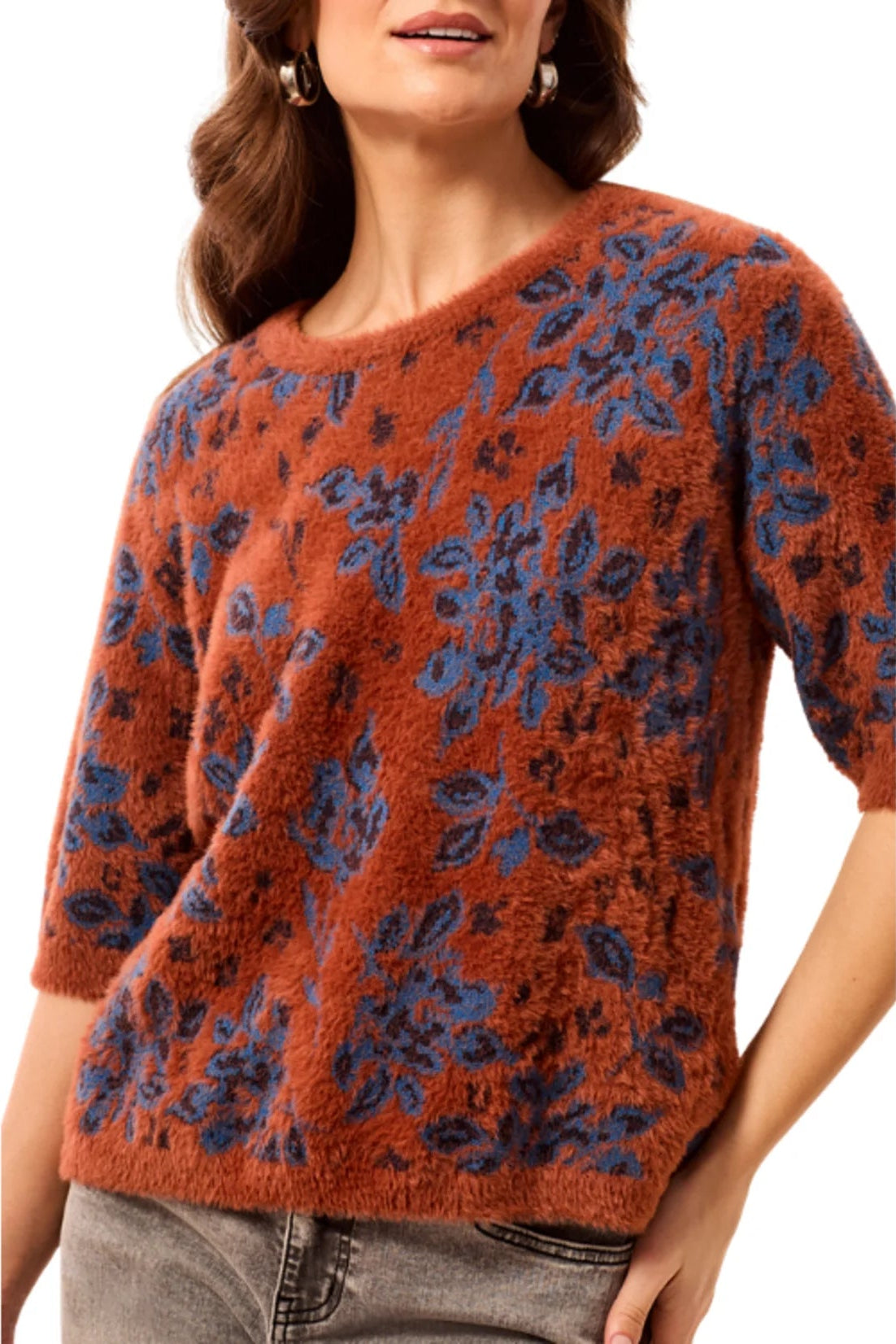 A woman wearing a fuzzy short-sleeve knit sweater with a rust-orange base and a blue floral pattern, paired with gray jeans against a plain white background.
