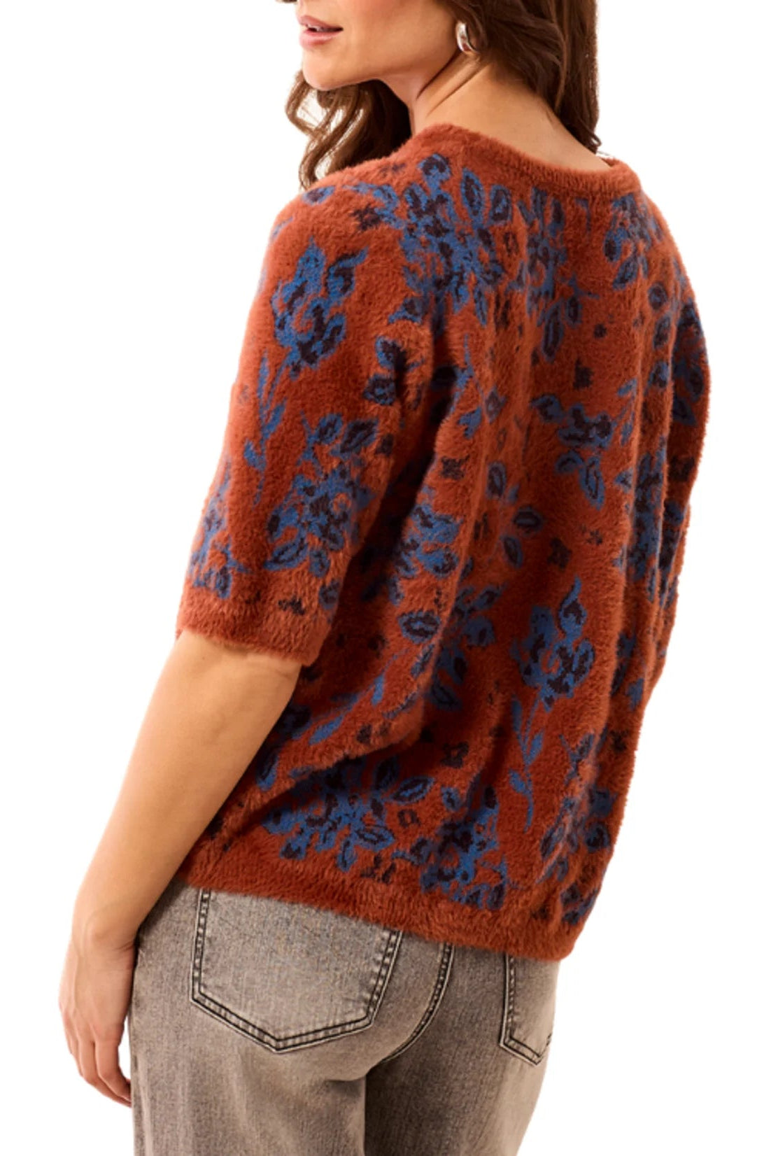 A woman wearing a fuzzy short-sleeve knit sweater with a rust-orange base and blue floral pattern, paired with gray jeans, shown from a side-back angle against a plain white background.