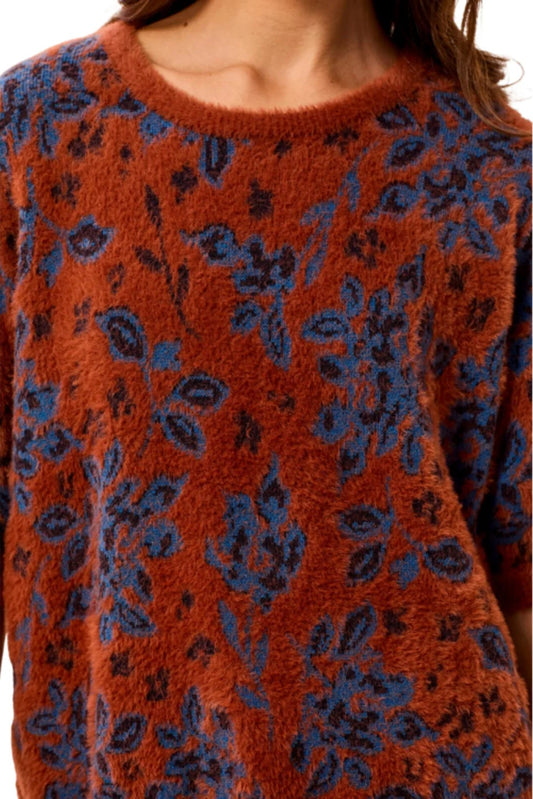 A close-up of a person wearing a fuzzy knit top with a rust orange base and a dark blue floral pattern, shown against a plain white background.