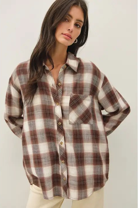 A woman wearing an oversized brown, beige, and white plaid button-down shirt, paired with light beige pants, against a plain light gray background.