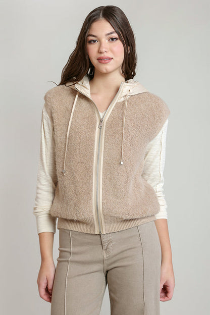 A woman modeling a beige fuzzy zip-up vest with a white hood and white long sleeves, paired with beige pants, against a light gray studio background.
