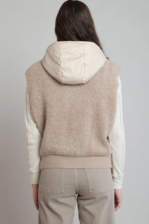 A model shown from the back wearing a light gray sleeveless knit hoodie vest over a cream ribbed long-sleeve top and beige trousers, against a neutral light gray background.