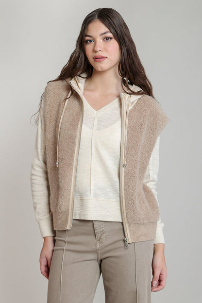 A woman modeling a beige fuzzy zip-front vest layered over a cream V-neck sweater and paired with taupe pants, photographed against a plain light gray studio background.