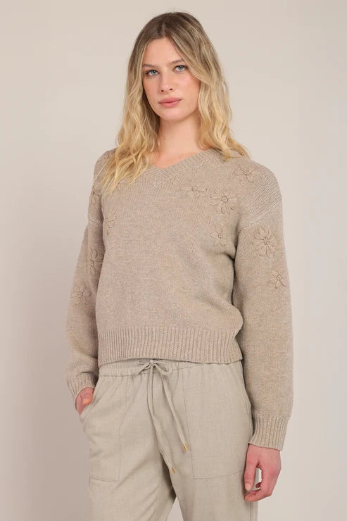 A woman modeling a light beige knit sweater with subtle floral embroidery on the sleeves, paired with matching beige drawstring trousers, against a plain light beige studio background.