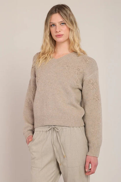A woman modeling a light beige knit sweater with subtle floral embroidery on the sleeves, paired with matching beige drawstring trousers, against a plain light beige studio background.
