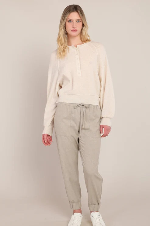 A woman modeling a cream-colored long-sleeve knit top and light beige jogger pants with white sneakers, standing against a solid light beige studio background.