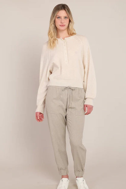 A woman modeling a cream-colored long-sleeve knit top and light beige jogger pants with white sneakers, standing against a solid light beige studio background.