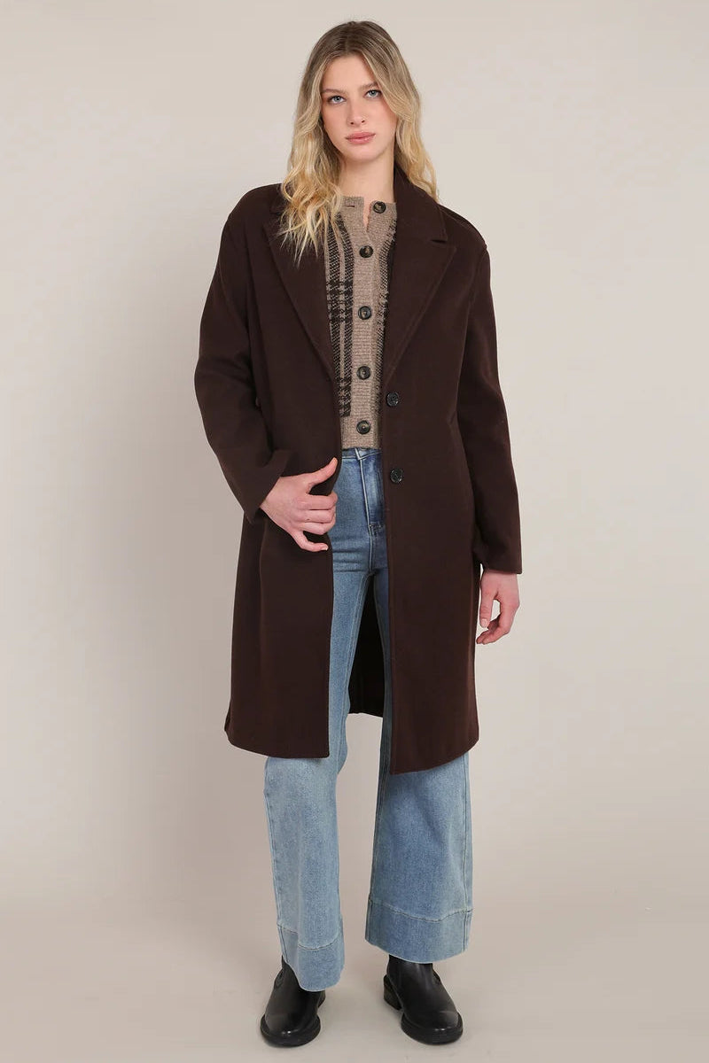 A woman modeling a long dark brown wool-blend coat over a beige plaid sweater, light blue wide-leg jeans, and black boots, photographed against a plain beige studio background.