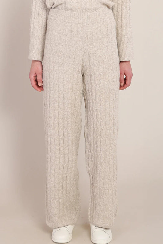 A person wearing cream-colored, cable-knit wide-leg pants and a matching sweater, paired with white sneakers, against a solid beige studio background.
