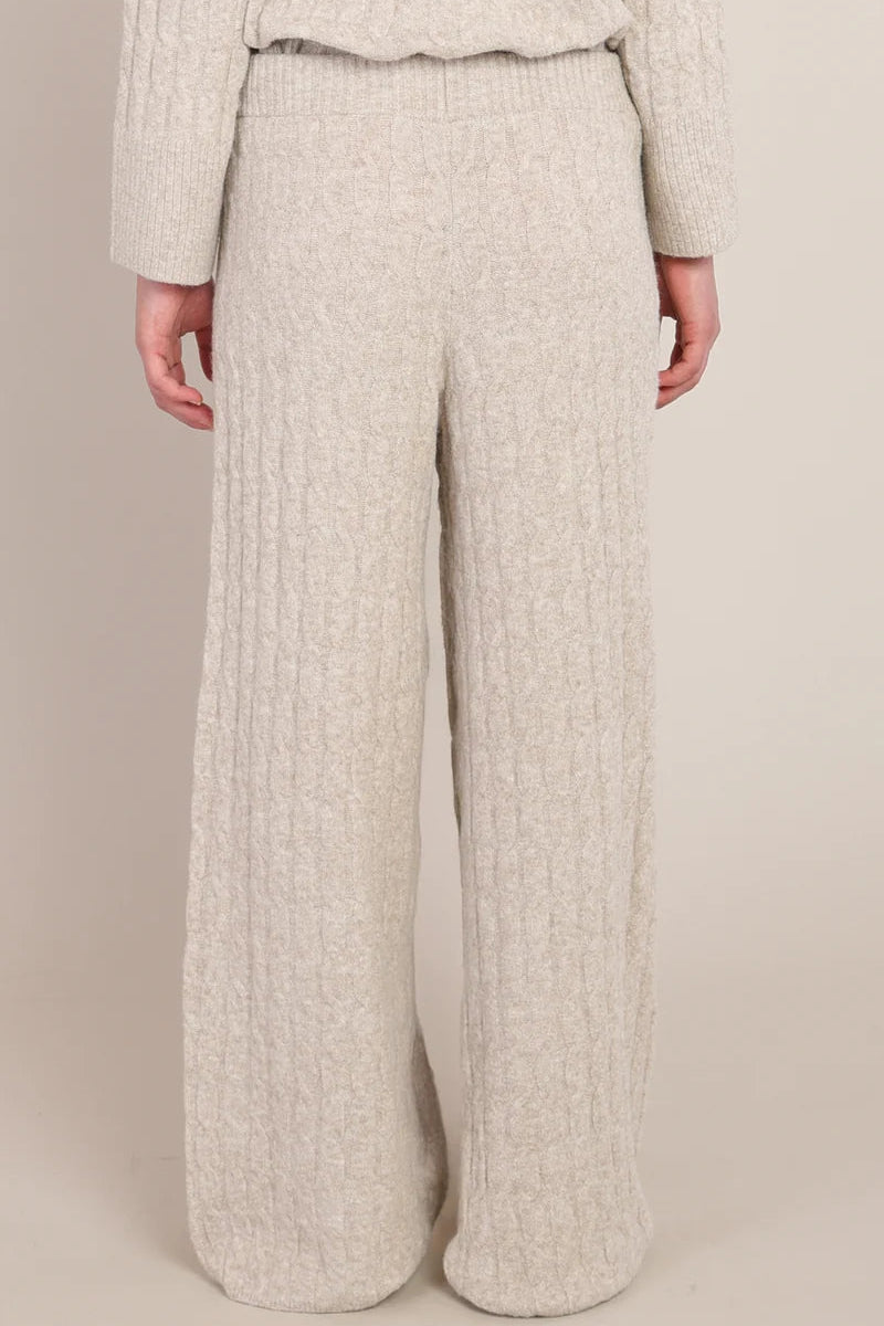 A person wearing a cream-colored, chunky cable-knit sweater and matching wide-leg knit pants, shown from the back against a solid beige studio background.