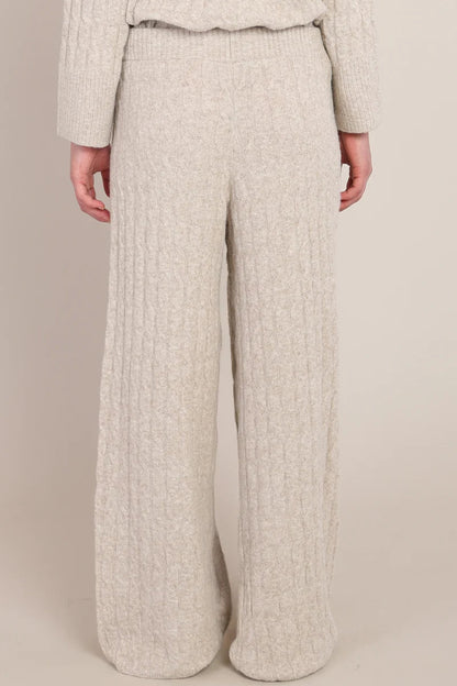 A person wearing a cream-colored, chunky cable-knit sweater and matching wide-leg knit pants, shown from the back against a solid beige studio background.