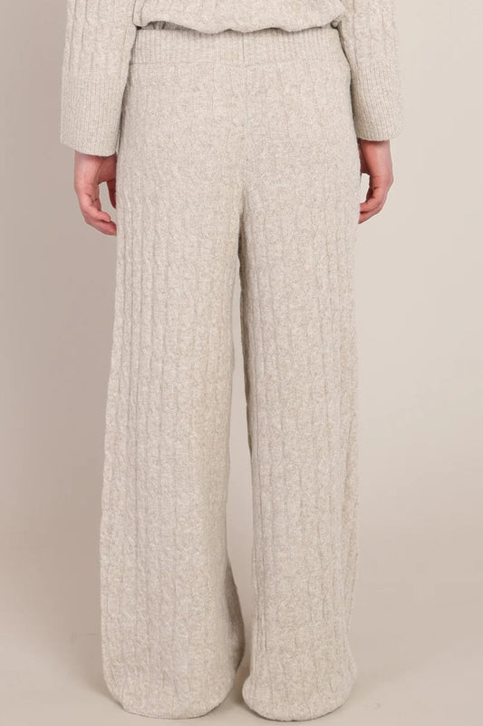A person wearing a cream-colored, chunky cable-knit sweater and matching wide-leg knit pants, shown from the back against a solid beige studio background.