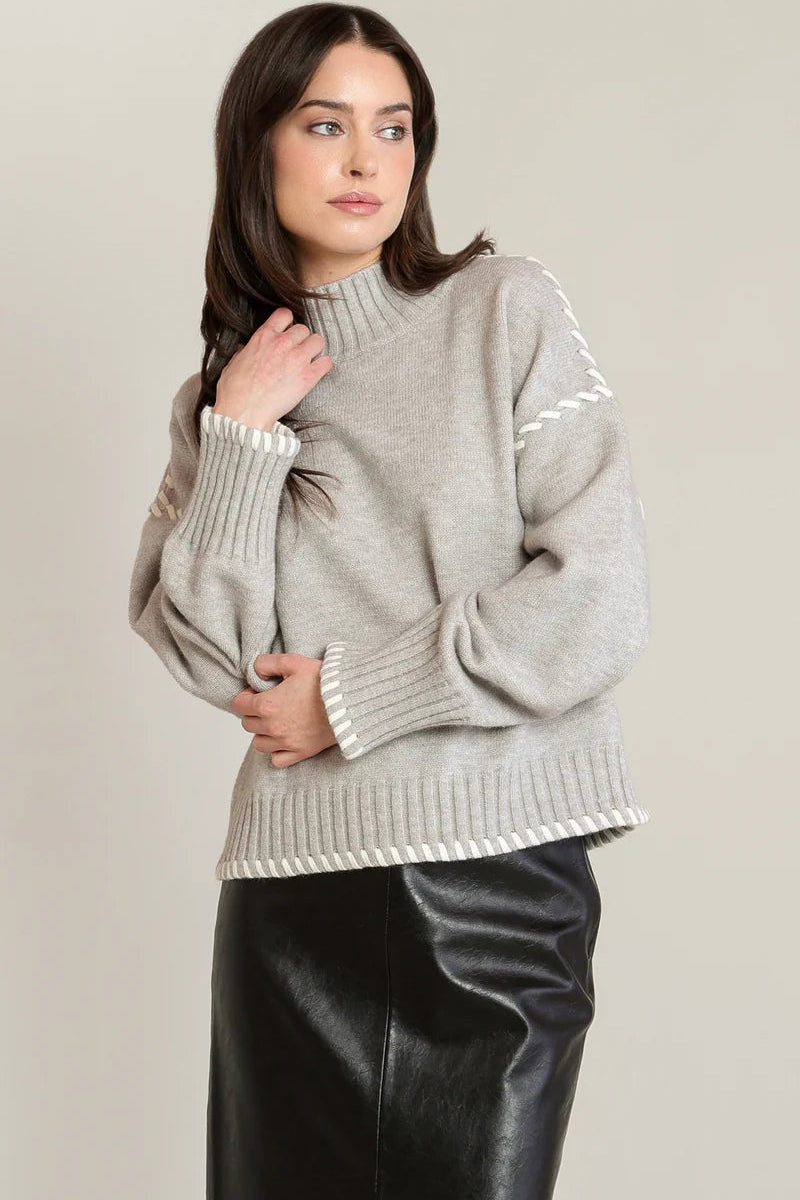 A woman wearing a light gray knit mock-neck sweater with white contrast trim and a black faux-leather skirt, standing against a plain light beige studio background.